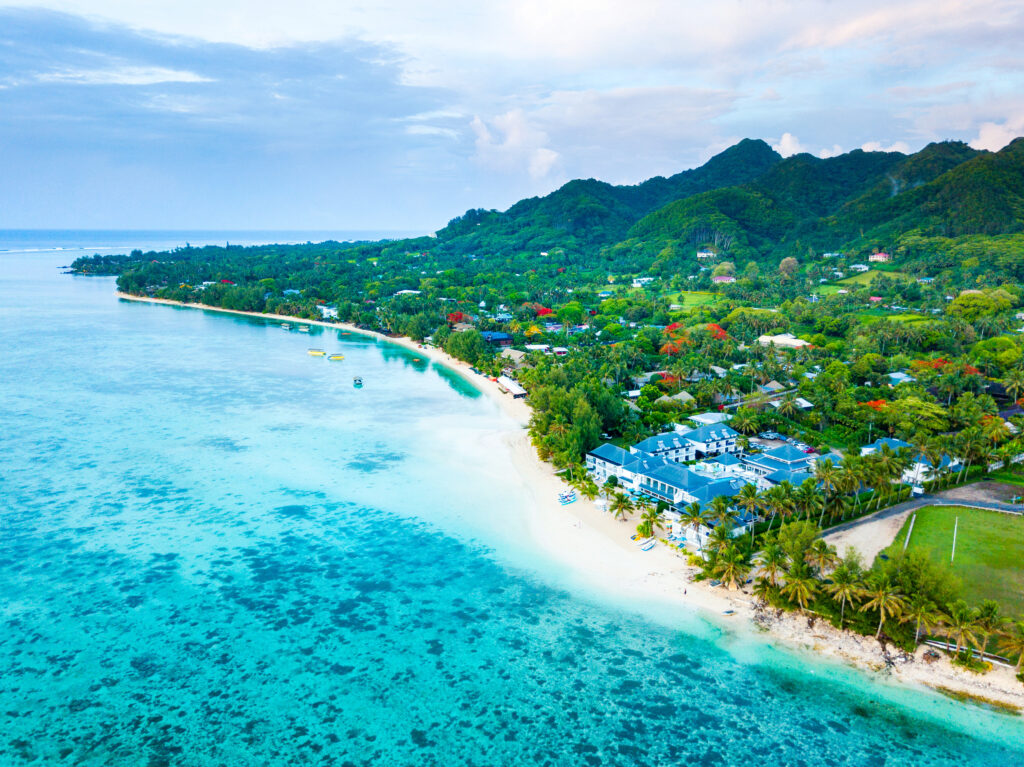 An aerial view of the coast of Rarotonga. It shows one of the small resorts and several colorful buildings along with the beautiful turquoise waters. 